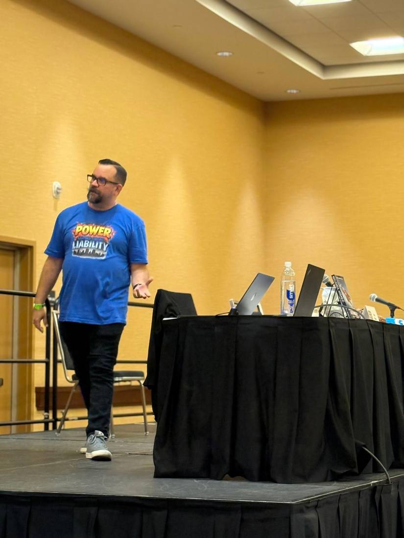 Brian Brijbag on stage at a comic convention panel, wearing a With Great Power Comes Great Liability shirt